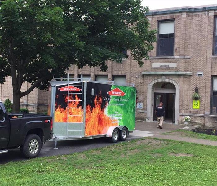 Black pickup truck with SERVPRO Job Trailer attached in front of a large brick school providing emergency services.