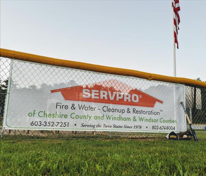 Company banner hanging from a chain link fence with a baseball cap and bat leaning up against it at a little league field